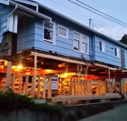 Blue house lift on piers. Evening. Lights on under house.