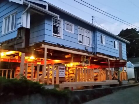 Blue house lifted on piers. Evening. Lights on under house.